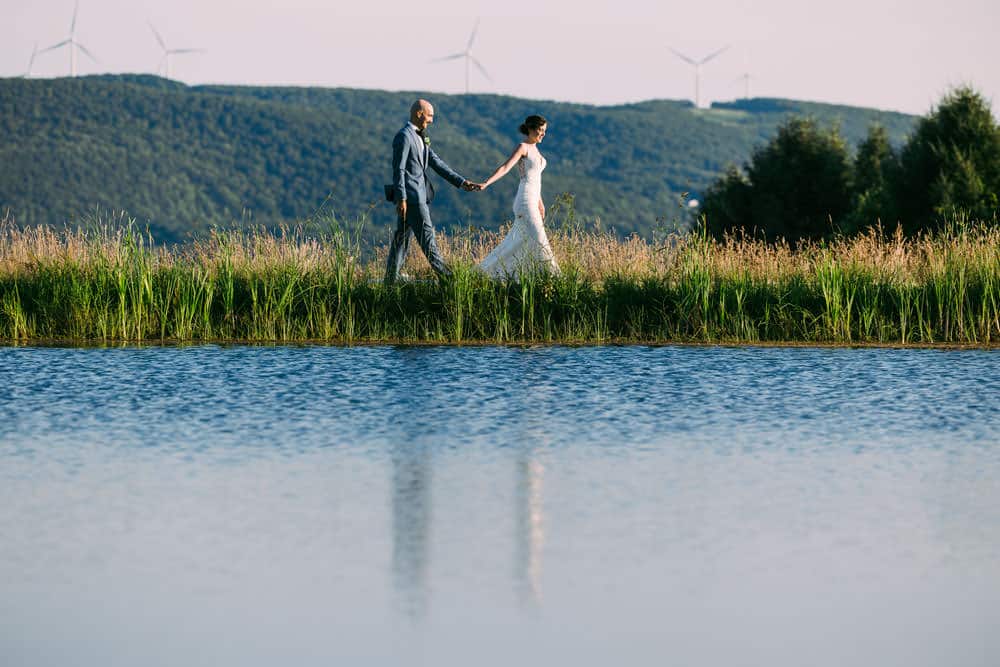 bride and groom walking with near the pond at Wren's Roost Barn and the hills of Naples, NY in the background